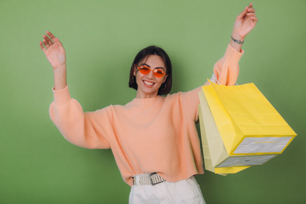 兴奋穿着休闲桃色毛衣的年轻女子被隔离在绿橄榄色的墙上 手里拿着时尚的购物袋 戴着橙色眼镜 跳跃着有趣的复制空间顾客女人买家