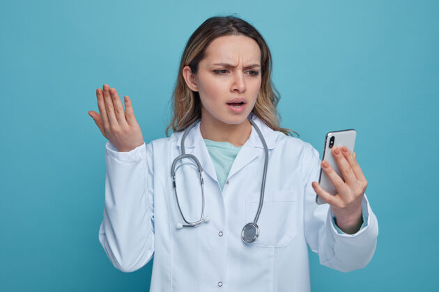 蓝色恼火的年轻女医生穿着医用长袍 脖子上戴着听诊器 手举着手机 看着手机穿手听诊器