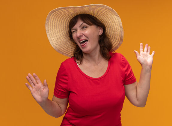 中等身穿红色t恤 头戴夏帽的中年妇女高举双臂站在橘色的墙上 脸上洋溢着幸福快乐的笑容年龄帽子身高