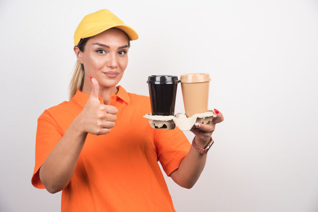 外卖金发女信使拿着两杯咖啡 在白墙上竖起大拇指快递女人塑料杯