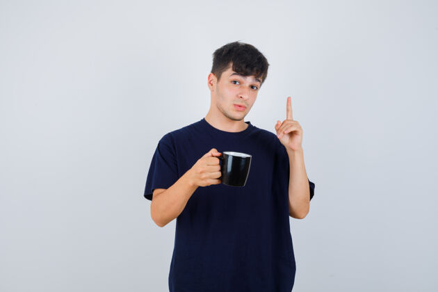 朋友年轻人的肖像显示尤里卡手势 指向上 拿着一杯饮料在黑色t恤衫和看起来聪明的前视图聪明成人肖像
