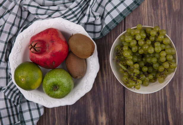 木头顶视图绿色葡萄碗石榴苹果猕猴桃和橘子在绿色方格毛巾木墙上石榴水果风景