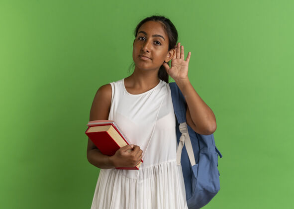 绿色年轻的女学生背着书包拿着带笔记本的书 表现出倾听的姿态抱着学生手势