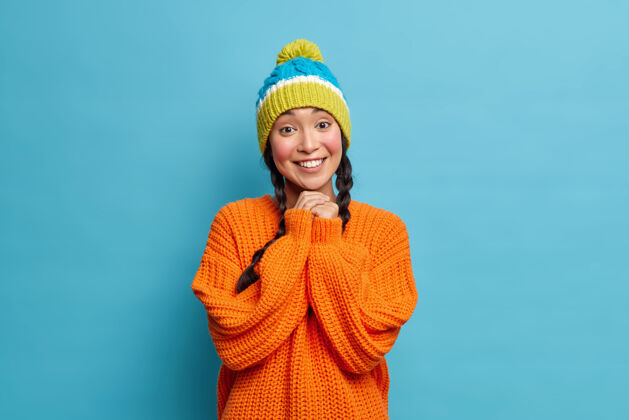 休闲美丽的亚洲女人的肖像 带着牙齿的微笑 双手合十 心情愉快 戴着冬天的针织帽子 穿着橙色的毛衣 在蓝色的墙上摆姿势女士可爱积极