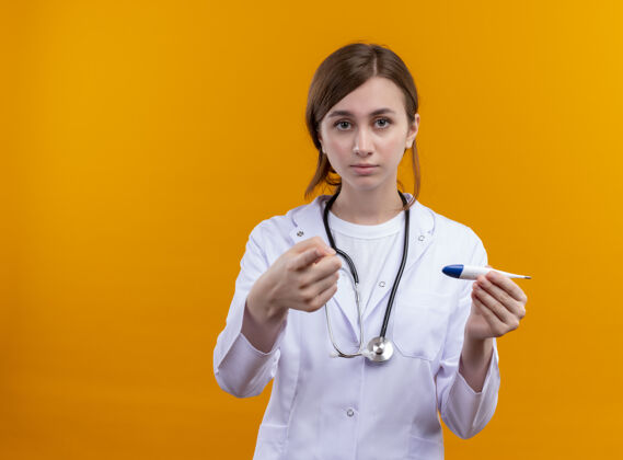 女人体贴的年轻女医生 穿着医用长袍 戴着听诊器 用手指举着温度计 站在隔离的橙色墙上 留着复印空间女人长袍拿着