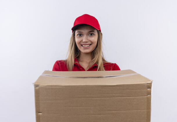 年轻微笑着的年轻送货女孩穿着红色制服 戴着帽子 手里拿着一个大箱子 孤零零地站在白色的墙上制服穿微笑