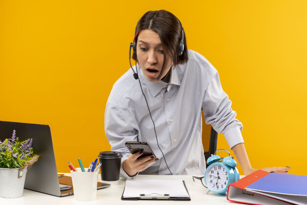 通话印象深刻的年轻呼叫中心女孩戴着耳机站在办公桌上拿着手机看橙色孤立居中办公桌年轻