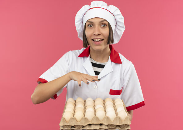 制服穿着厨师制服的年轻女厨师手拿一盒鸡蛋 手放在鸡蛋上方 与粉红色的鸡蛋隔离开来年轻印象鸡蛋
