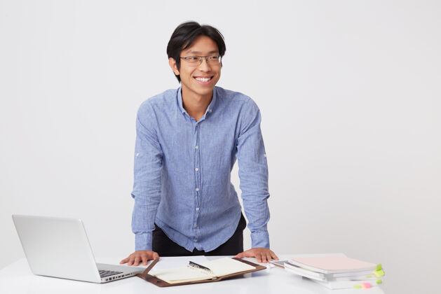 欢呼快乐自信的亚洲年轻商人戴着眼镜站在桌边工作 笔记本电脑和笔记本电脑隔着白墙站立规划师离开