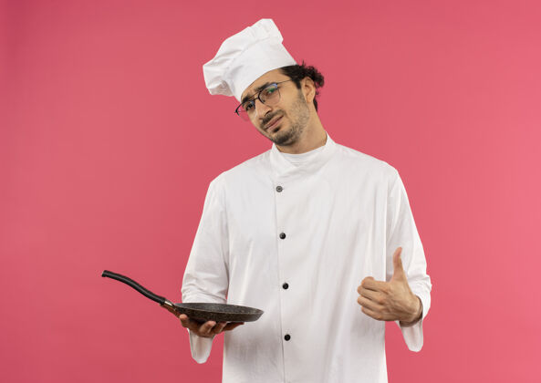 持有穿着厨师制服戴着眼镜拿着煎锅的年轻男厨师把大拇指孤立地竖在粉红色的墙上不愉快平底锅大拇指