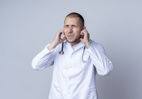 脖子恼怒的年轻男医生穿着医用长袍 脖子上戴着听诊器 把手指放在耳朵上 闭着眼睛 隔离在白色的空间里耳朵复制封闭