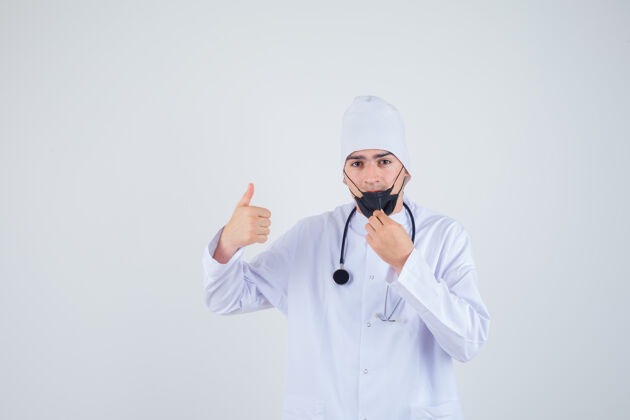 年轻年轻人试着拿起口罩 穿着白色制服 戴着口罩 大拇指朝上 看上去很满意正视图信心男性医生