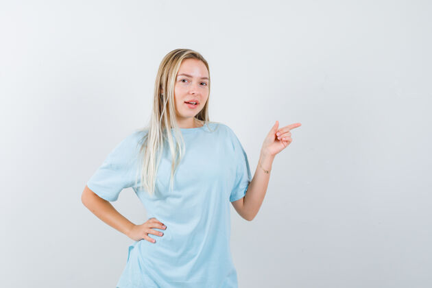 目录金发女孩用食指指着右边 手放在腰上 穿着蓝色t恤 看起来很漂亮 正面视图肖像时尚指向