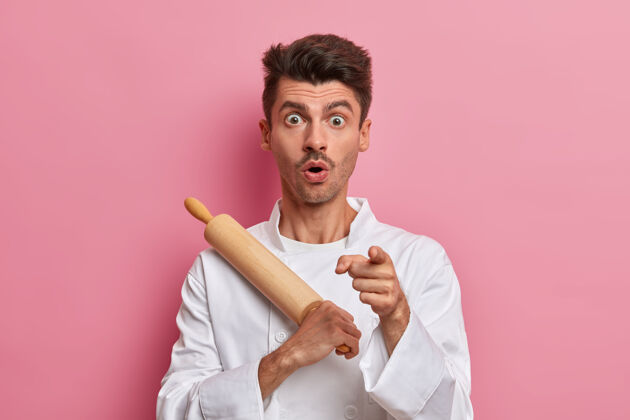 害怕震惊的男面包师拿着擀面杖指着前面 准备做饭 在厨房工作 穿制服举行男人准备