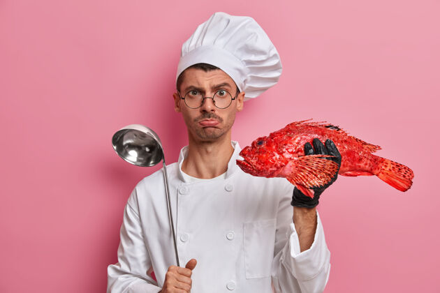 餐厅严肃的厨师拿着勺子和鲈鱼 展示鱼汤的烹调过程 在餐厅准备聚会 戴着圆眼镜 穿着白色制服室内厨具人