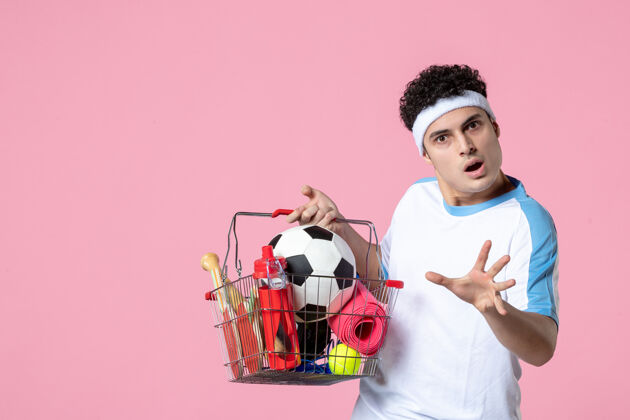 购物正面图穿着运动服的年轻男子 带着装满运动用品的篮子 粉色的墙壁年轻的男性人篮子