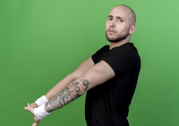 墙自信的年轻运动型男子戴着腕带伸展双臂隔离在绿墙上年轻男人运动