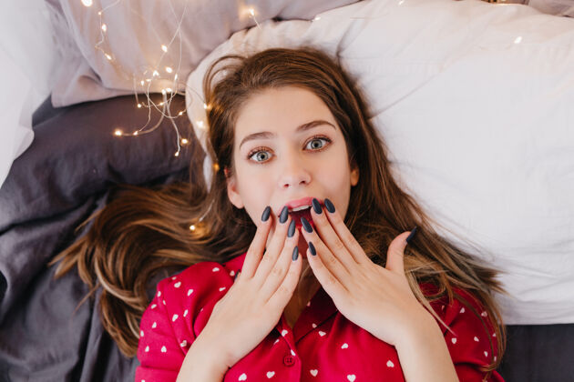 肖像可爱的黑发女孩 黑指甲 躺在床上 表示惊讶头顶照片蓝眼睛无忧无虑的年轻女子穿着红色睡衣床睡觉睡衣