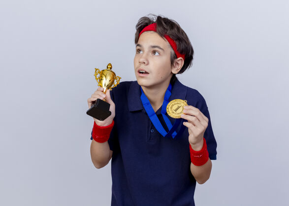 站着惊讶的年轻英俊的运动男孩戴着头带和护腕 戴着牙套 脖子上戴着奖牌 手里拿着奖牌和优胜者杯 看着隔离在白墙上的一面感觉人腕带