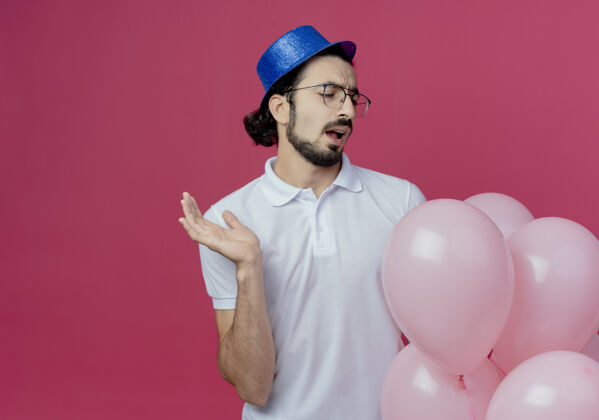 粉色一个戴着眼镜和蓝帽子的帅哥拿着气球 看着气球 摊开手孤立在粉红色的上面帅气帽子戴着