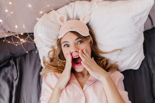 积极戴着粉红色眼罩躺在枕头上的一个令人惊奇的迷人的年轻女子头顶上的金发女孩的画像 早上在床上摆姿势年轻快乐表情