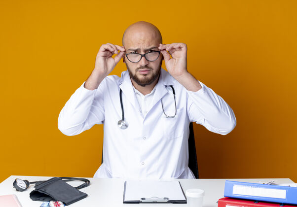 听诊器严格年轻的秃头男医生穿着医用长袍和听诊器坐在工作台上拿着医疗工具戴上橙色的眼镜工作医生长袍
