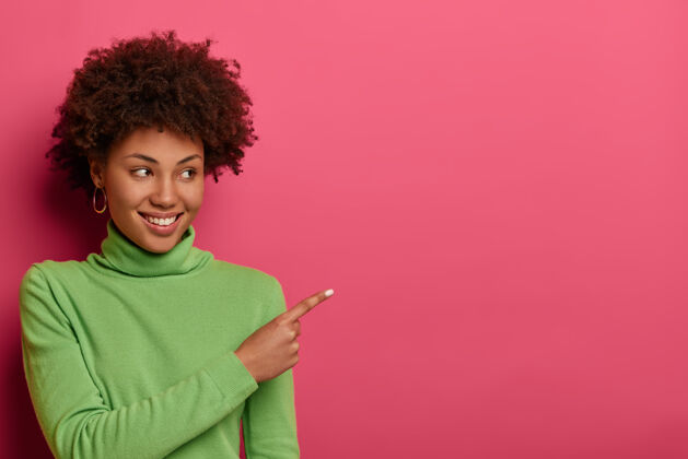 青少年笑容可掬的卷发女子横拍在自由空间标明 为您的广告展示场所 吸引眼球销售 穿着绿色高领毛衣 孤立在充满活力的粉色墙上罗西毛衣站立