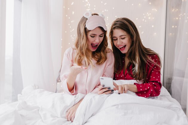 室内穿着时尚睡衣的漂亮女孩在发短信迷人的年轻女士一起坐在床上看智能手机屏幕的室内写真年轻快乐电话
