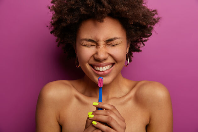 牙齿无上装快乐的非洲女人用牙刷保持口腔卫生 笑容灿烂 拥有洁白完美的牙齿 斜视着脸 对着紫色的墙壁摆姿势牙齿美白 口腔美容 牙齿护理年轻美白种族