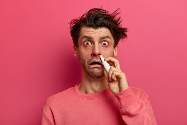 人震惊的男人有红肿的眼睛 溅鼻涕 治疗过敏性鼻炎 有家庭治疗 盯着粉红色的墙壁摆姿势 在里面滴药感冒或过敏的症状恢复不安治疗