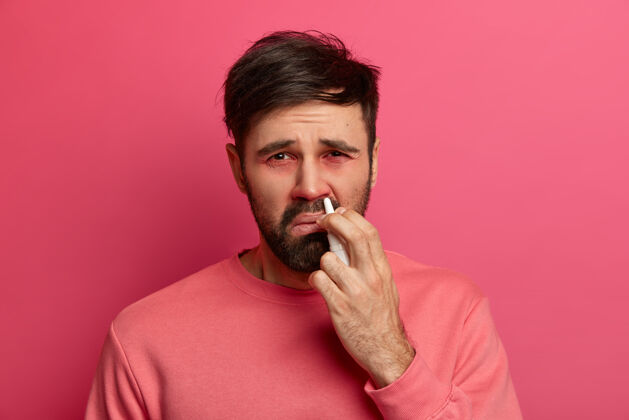 人心烦意乱的病人在鼻子里喷过敏药 感冒 鼻炎 眼睛红肿 穿着休闲服 对着粉红色的墙壁摆姿势疾病治疗理念疾病不高兴治疗