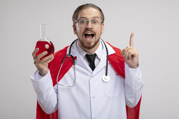 瓶子年轻的超级英雄穿着医用长袍 戴着听诊器 戴着眼镜 手里拿着装满红色液体的化学玻璃瓶 指着白色背景上的隔离墙年轻超级英雄化学