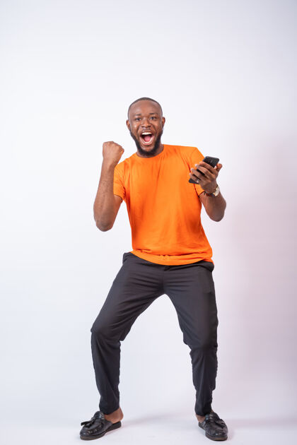 男人兴奋的尼日利亚年轻人一边庆祝 一边看着隔离在白墙上的手机穿着电话设备