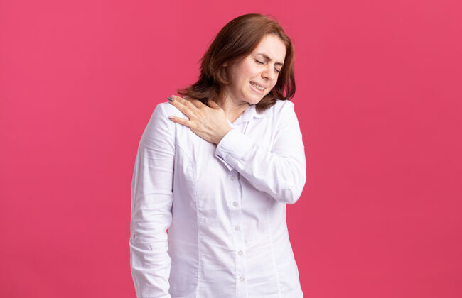 衬衫穿白衬衫的年轻女子看起来身体不适 站在粉红色的墙上摸着肩膀感到疼痛感觉女人肩膀