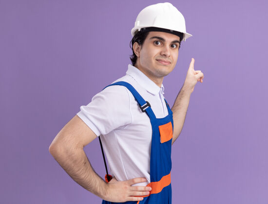 指点年轻的建筑工人穿着建筑制服 戴着安全帽 面带微笑 自信地用食指指着紫色墙壁上的东西自信制服目录