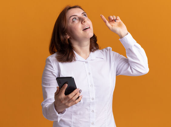 目录身穿白衬衫 手持智能手机 面带微笑的年轻女子站在橙色的墙上 食指有了新的想法衬衫显示女人