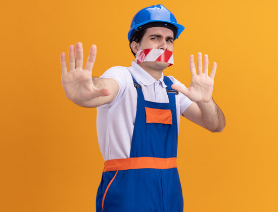 建筑年轻的建筑工人穿着建筑制服 戴着安全帽 嘴上缠着胶带 站在橘色的墙上 吓得伸出手来做防御手势胶带站着防御