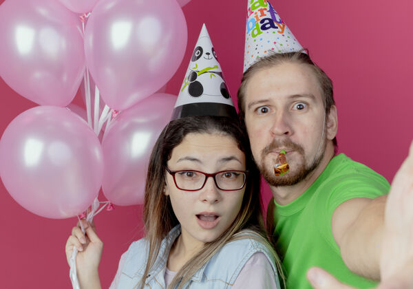 年轻这对戴着派对帽的年轻夫妇看起来很惊讶 女孩手里拿着氦气球 男人吹着哨子孤立地站在粉红色的墙上粉色女人男人