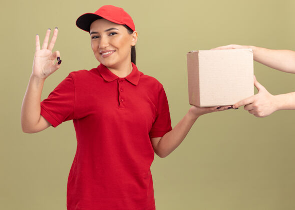 制服年轻的送货员身着红色制服 头戴鸭舌帽 微笑友好 站在绿色的墙壁上 一边接受包装箱 一边展示ok标志年轻送货朋友