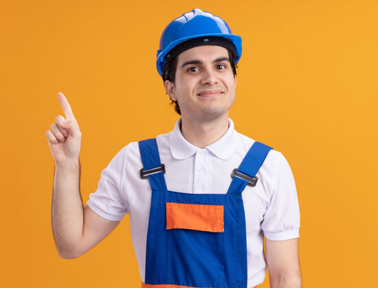 建筑工人年轻的建筑工人穿着建筑制服 戴着安全帽 微笑着自信地指着前面站在橙色的墙上的食指手指站立头盔