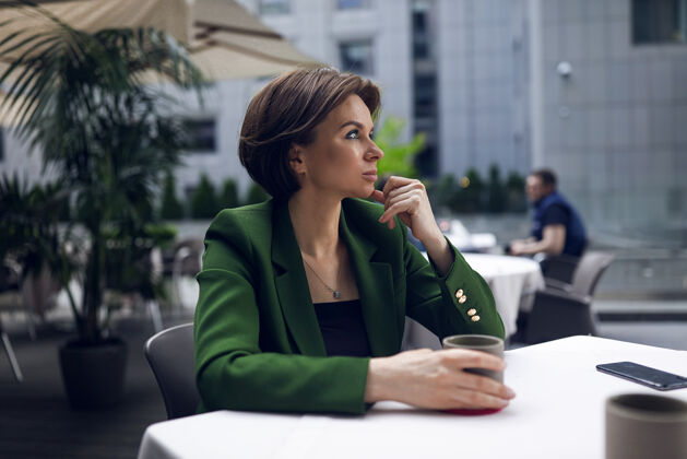 专业女商人坐在咖啡馆里 在所有的会议和面试后休息绿色时尚夹克和黑色衬衫 短发 裸妆一杯热咖啡杯子夹克办公桌