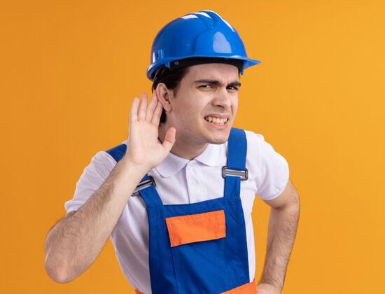八卦年轻的建筑工人穿着建筑制服 戴着安全帽 用手捂着耳朵看着前面 试图听站在橙色墙壁上的流言蜚语男人安全耳朵