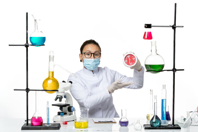时钟前视图：穿着医疗服的女化学家 戴着面罩 拿着浅白色背景上的时钟病毒实验室化学冠状病毒飞溅持有实验室视图