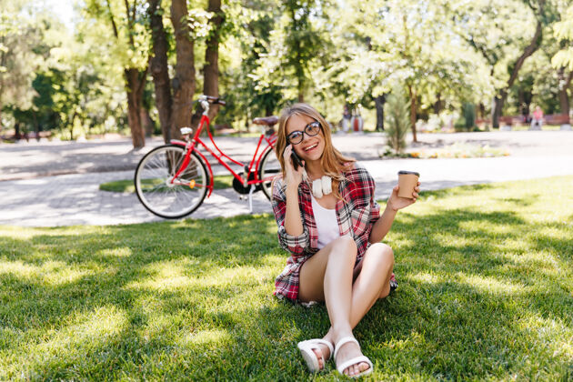 娱乐穿着夏装的快乐女人坐在草地上喝着咖啡戴着眼镜的迷人女孩在大自然里打电话的户外镜头人漂亮骑