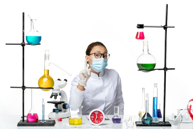 坐着前视图穿着医疗服的女化学家戴着面罩坐在浅白色的背景上 化学实验室的病毒冠状病毒-飞溅实验视图面具