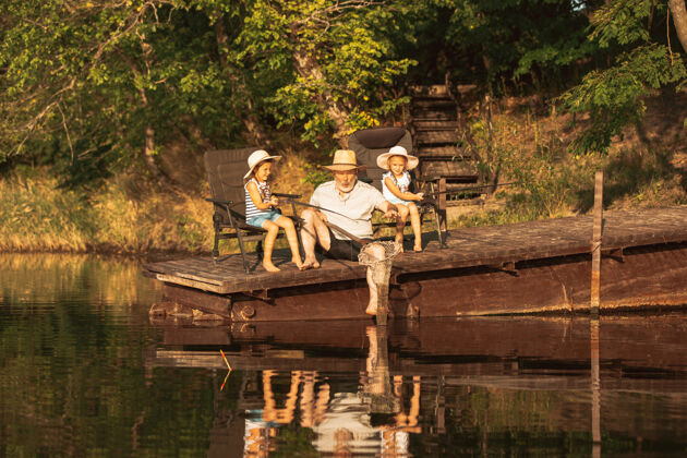 孩子可爱的小女孩和他们的爷爷在湖边或河边钓鱼在夏天的日落时分 在靠近水和森林的码头上休息家庭 娱乐 童年 自然的概念淡水童年爷爷