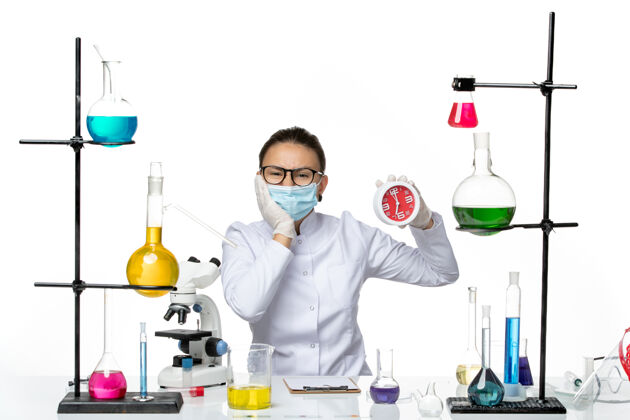 保持正面图白色办公桌上戴着面具的女化学家拿着时钟病毒实验室化学冠状病毒-飞溅面具喷溅外套