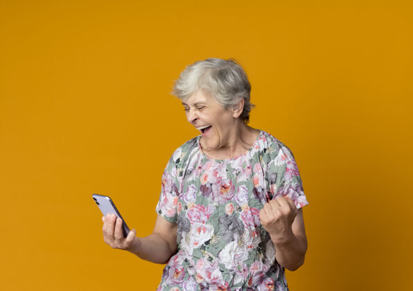 老人快乐的老妇人看着电话握着拳头孤立在橙色的墙上拳头抱着女人