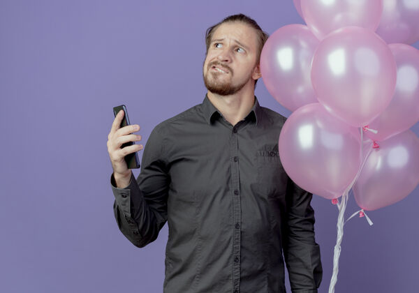 气球困惑的帅哥拿着氦气球站在紫色的墙上孤立地看着电话起来迷糊紫色