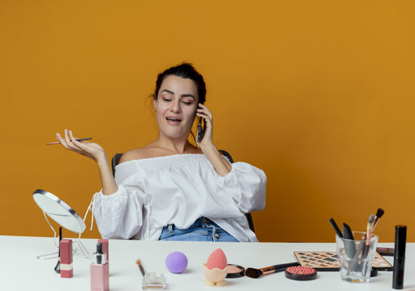 坐着美丽快乐的女孩坐在桌子旁 拿着化妆工具讲电话 拿着化妆刷隔离在橙色的墙上拿着橘子快乐